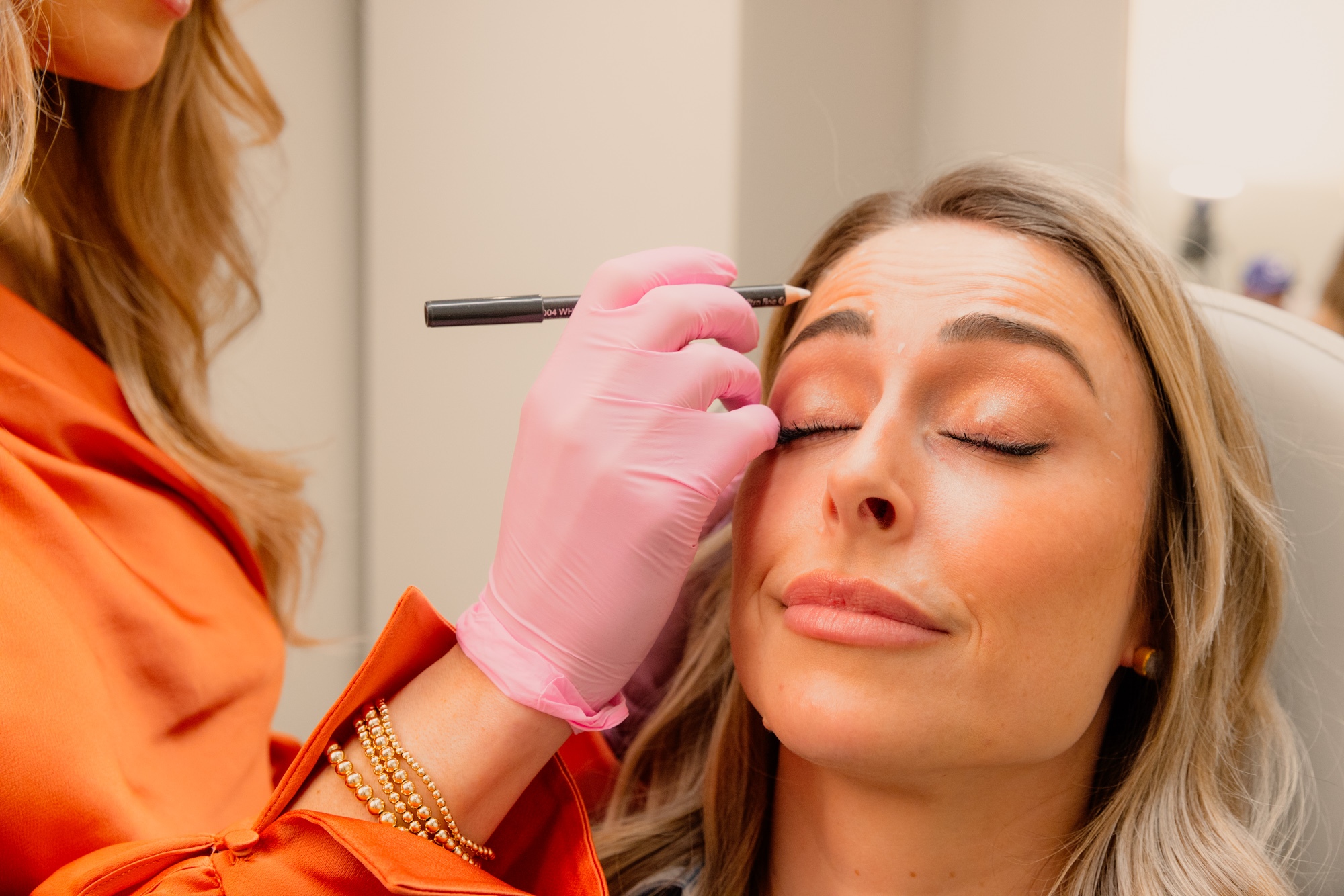 A female Artisan injector, wearing and orange blouse and pink gloves, marks injection sites on a female patient's face for dermal fillers. In Atlanta, Artisan specialists use dermal fillers, biostimulators and other cosmetic injectables for facial balancing.
