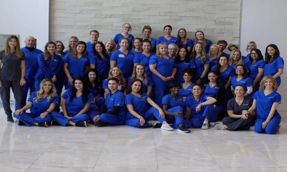 group photo of about 30 people wearing royal blue scrubs, smiling against grey marble floor and wall