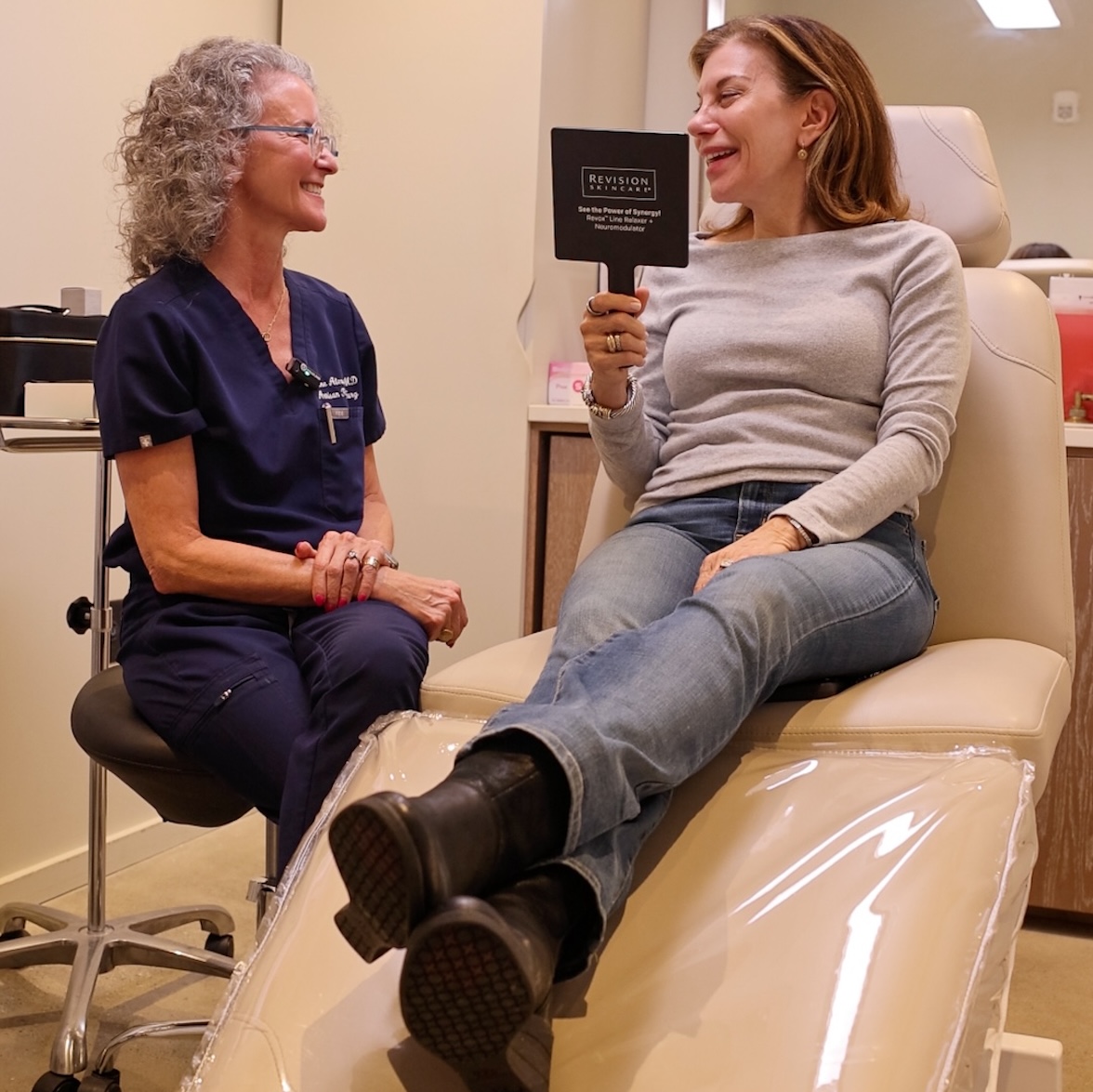 Dr. Alexander sits and laughs with a female patient who reclines on a treatment chair. The patient holds a mirror, happy with her blepharoplasty in Atlanta.